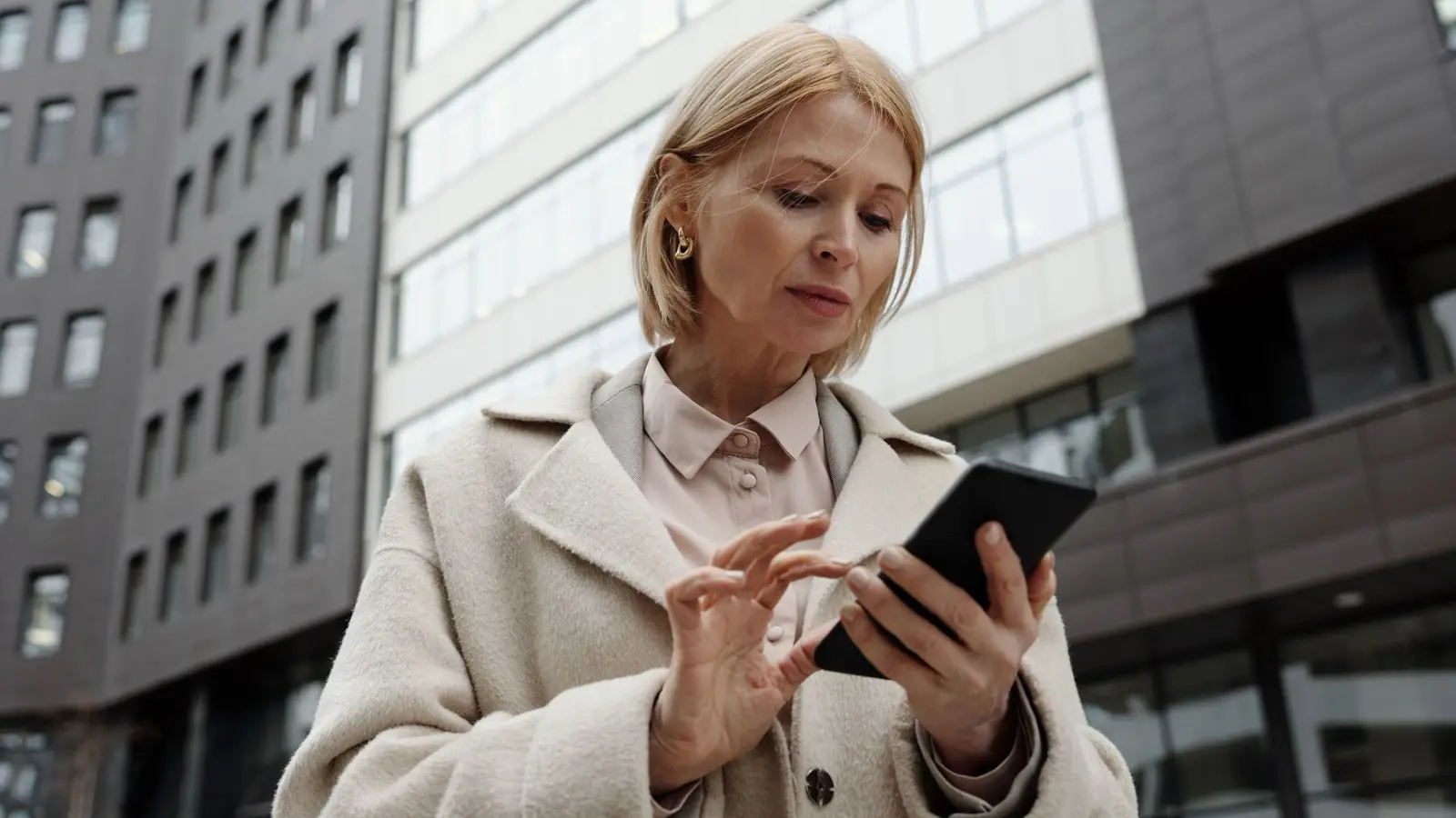 A woman in a light coat looks at a smartphone she holds in one hand, standing in front of office buildings.