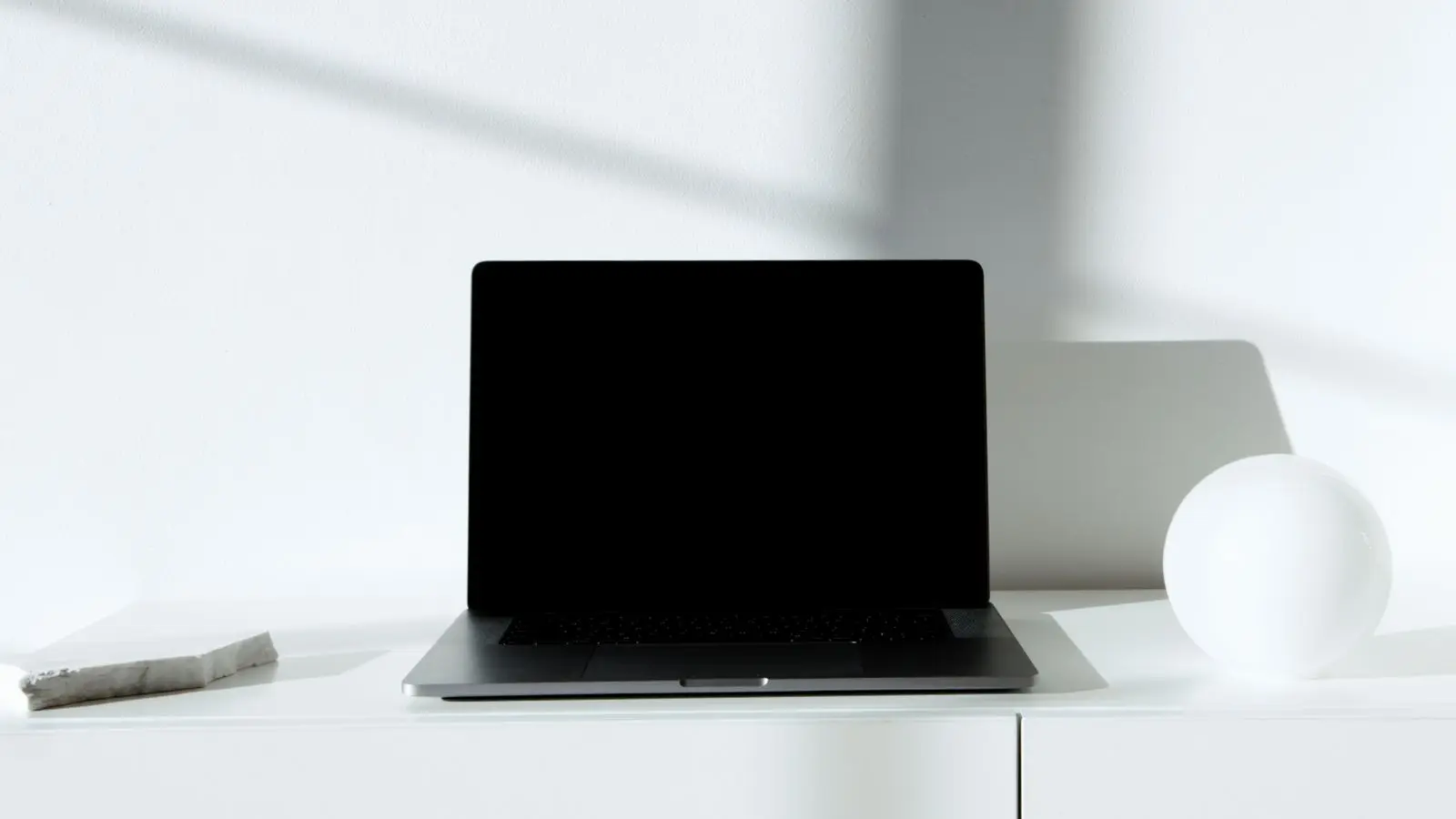 An open laptop with a dark screen on a white shelf, beside a white sphere and a stone tile.