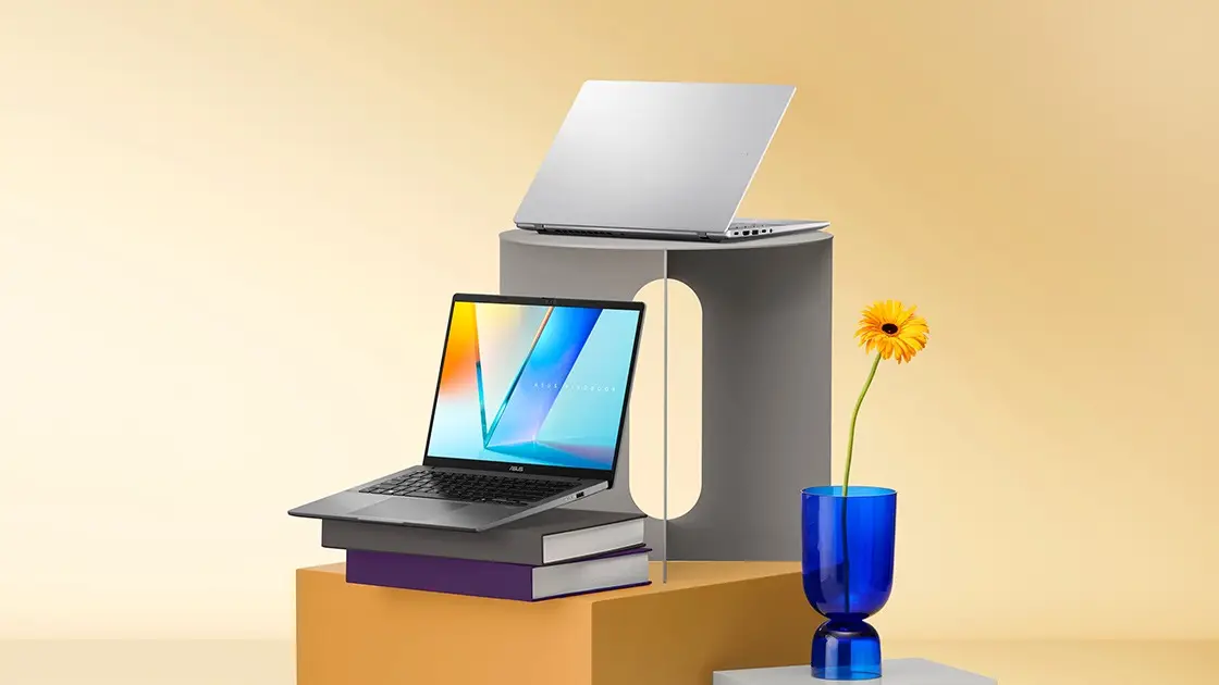 Two ASUS Vivobook S16 laptops on stacked books and a grey stand against a warm yellow backdrop, one open showing a blue and orange wallpaper, one silver lid-up, with a yellow flower in a blue vase.