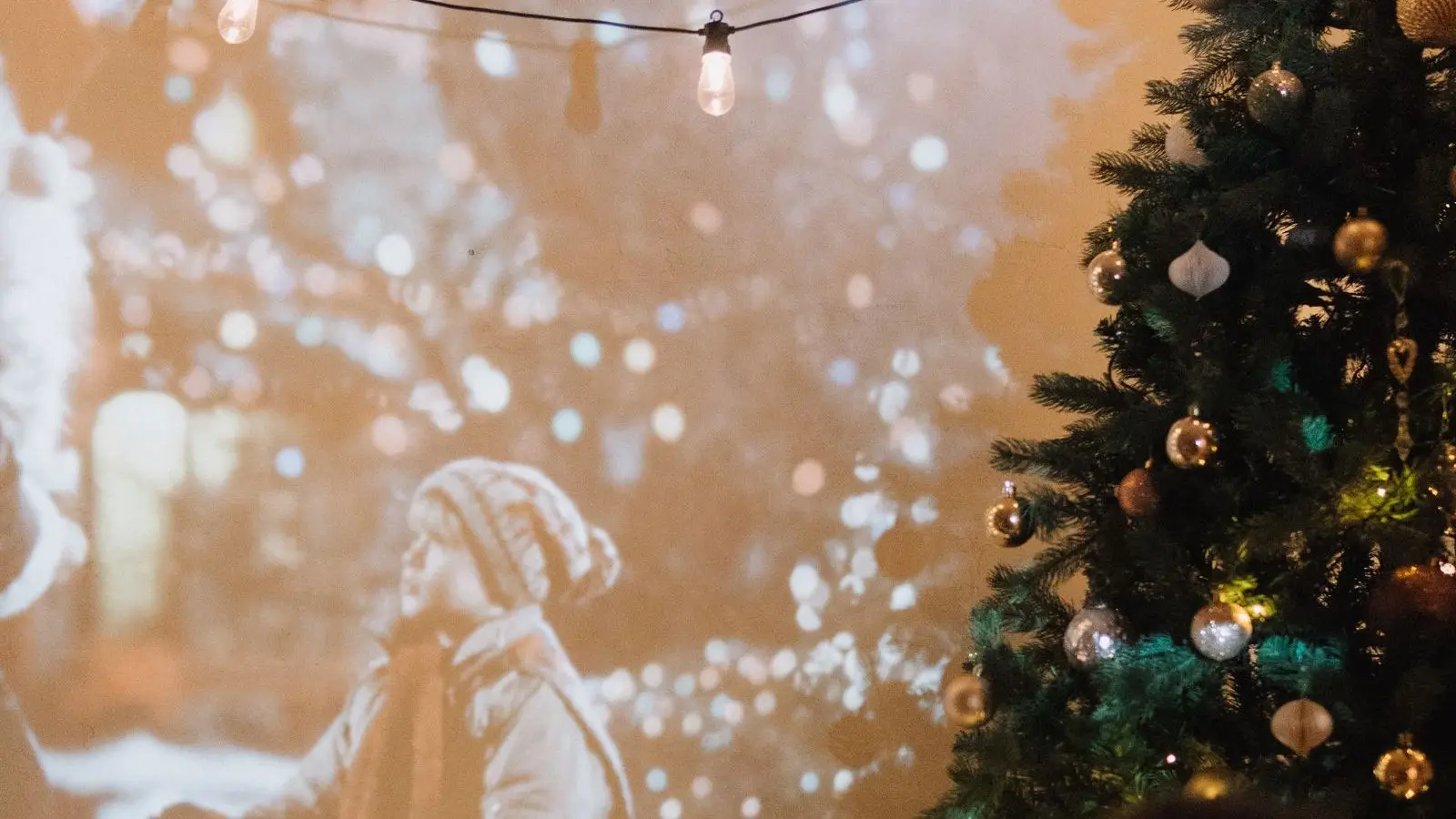 A film projected onto a wall in a dim room, with string lights and a decorated tree beside it.