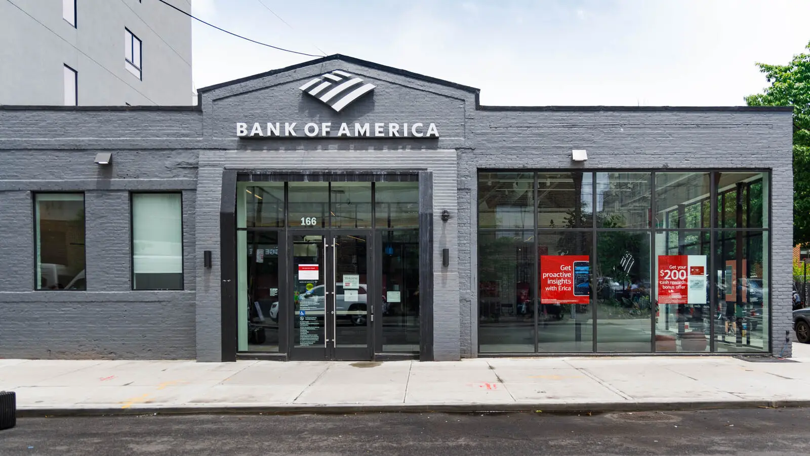 A Bank of America branch with a grey brick front, the bank's logo above the door and two red promotional posters in the window.