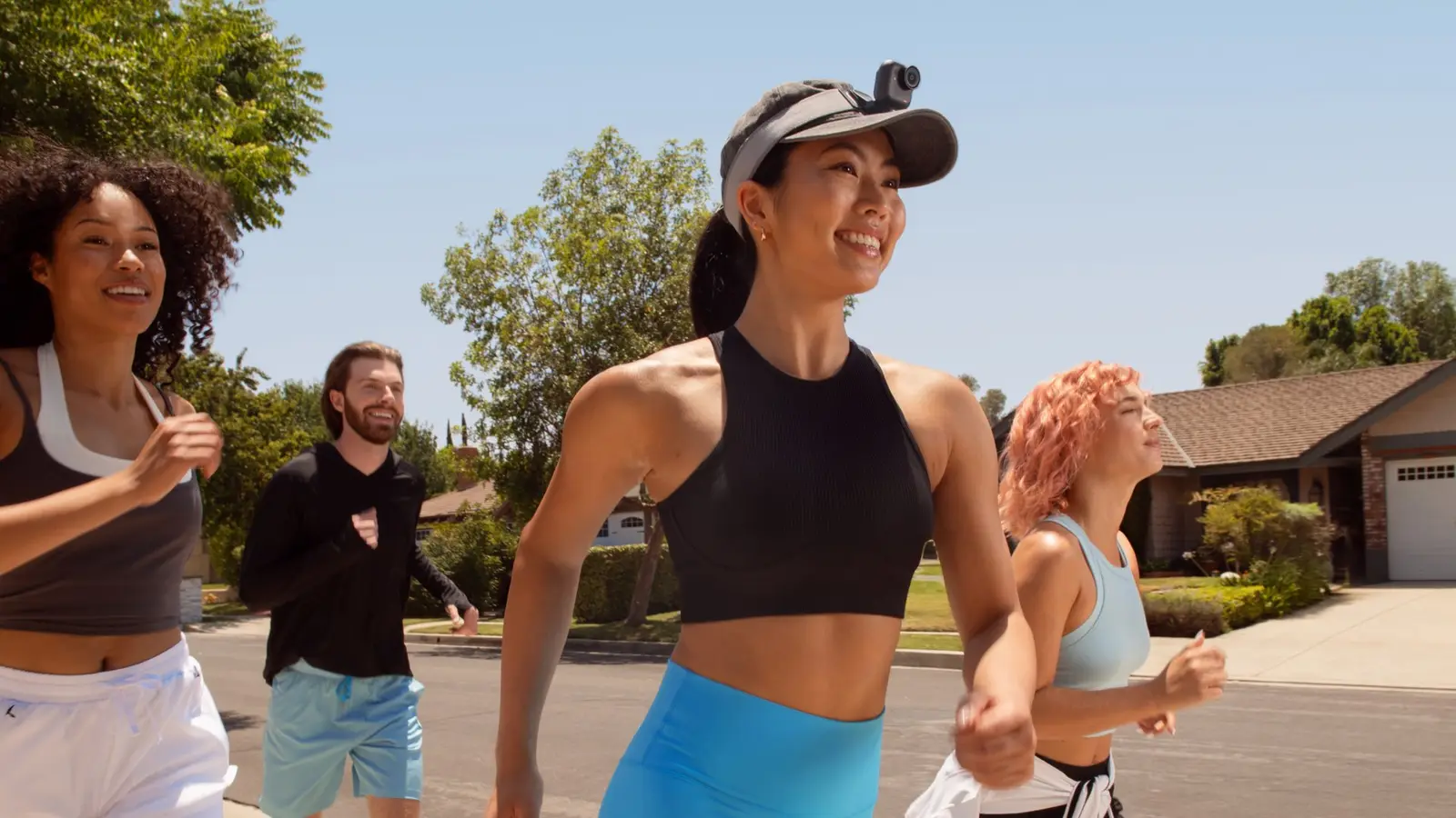 A smiling runner in the foreground with a small camera mounted on the front of the runner's cap, jogging with three other people on a residential street.