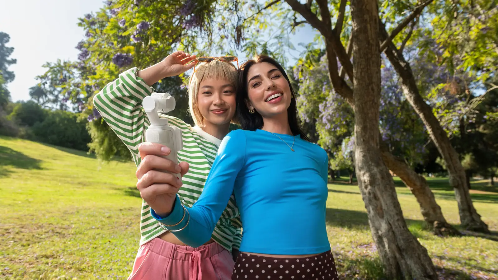 Two people standing close together in a park under a tree, one holding a small white handheld gimbal camera at arm's length.