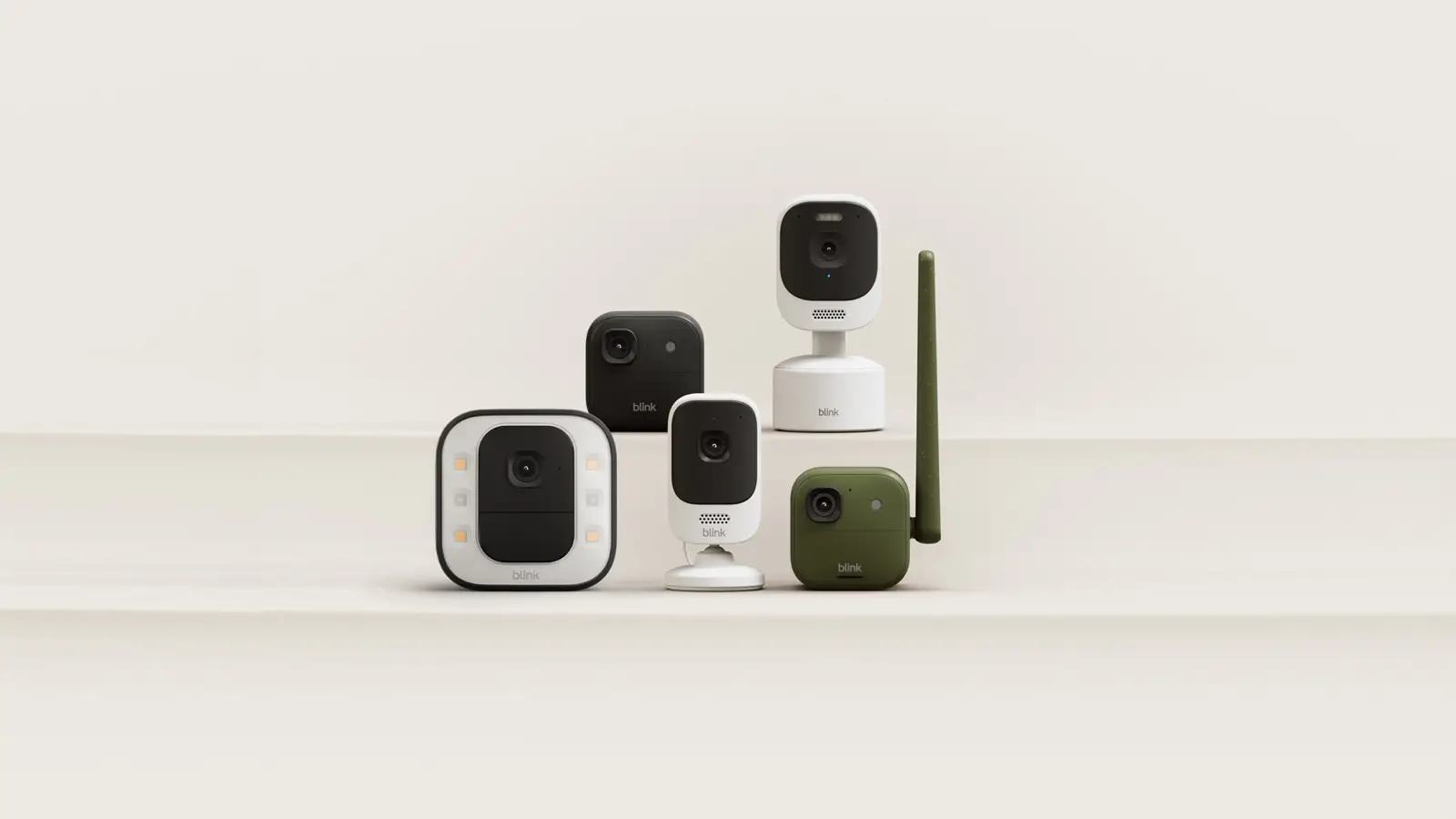Five new Blink cameras in white, black and olive green, one with a green antenna and one with a ring of lights, on a cream stepped background.
