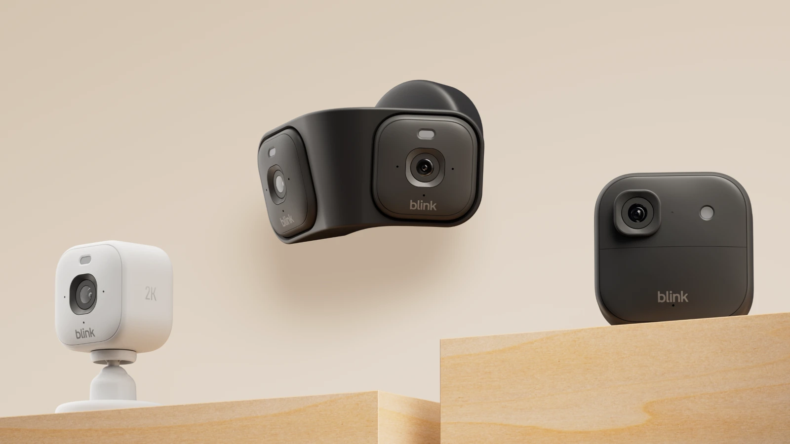 Three Blink cameras on wooden platforms against a beige wall: a white Mini 2K+ on a stand, a black twin-lens Arc in the middle and a black outdoor camera on the right.