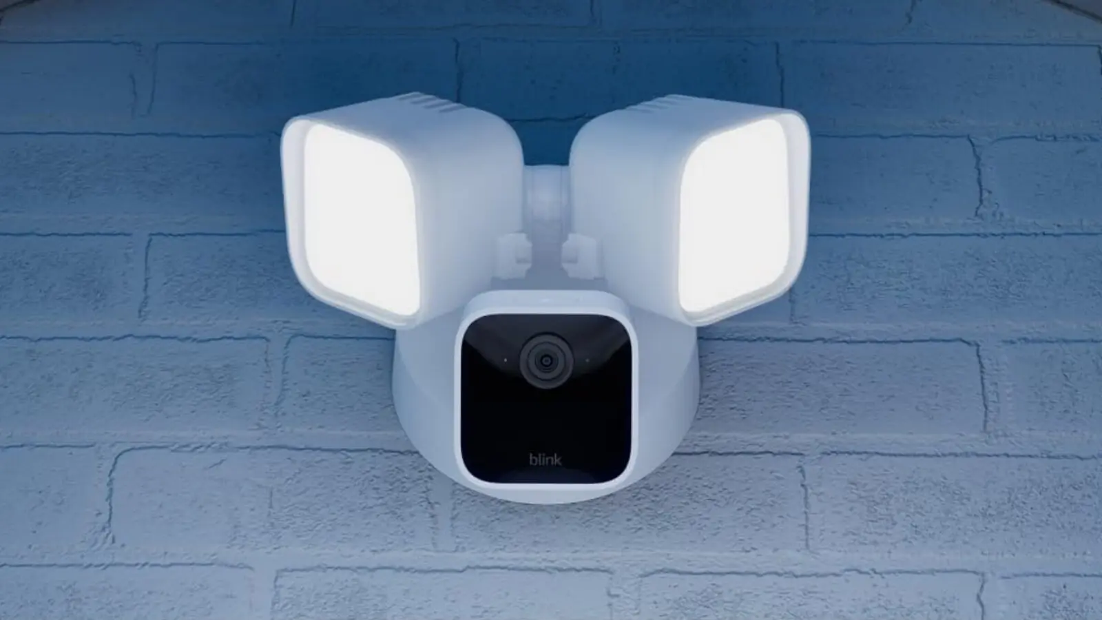 A white Blink Wired Floodlight Camera mounted on a blue-painted brick wall, with two lit floodlight heads above a camera with a black front.