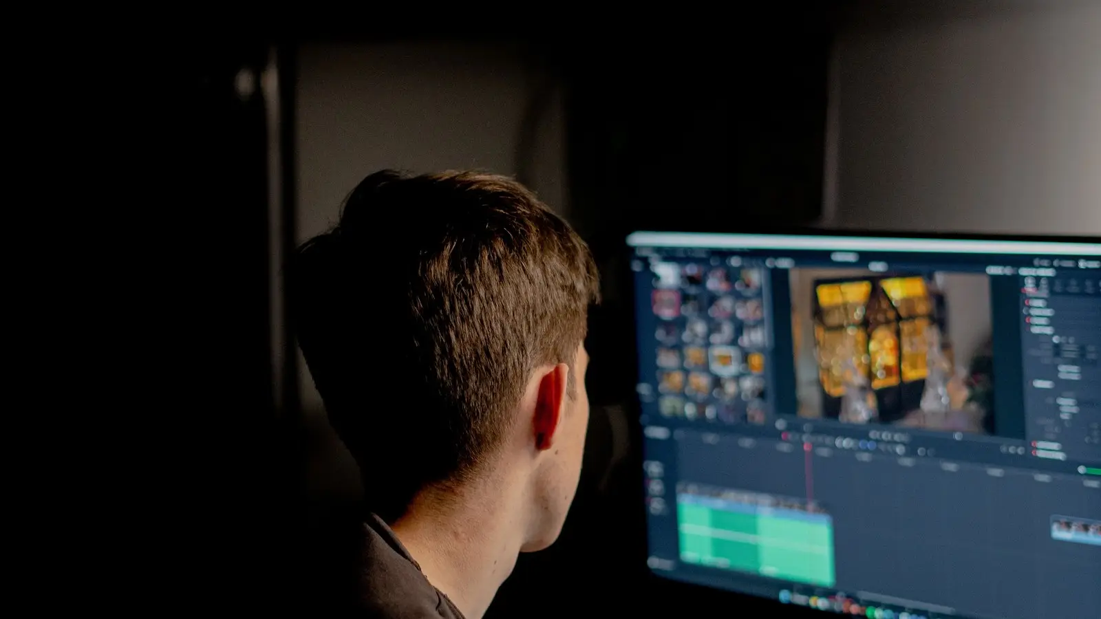 A man seen from behind watches a video editing timeline and preview window on a monitor in a dark room.