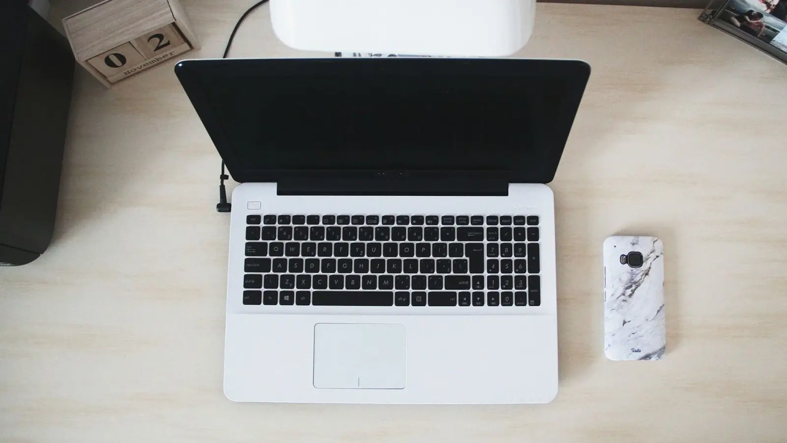 A white laptop seen from above on a pale wooden desk, with a phone in a marbled case beside it.