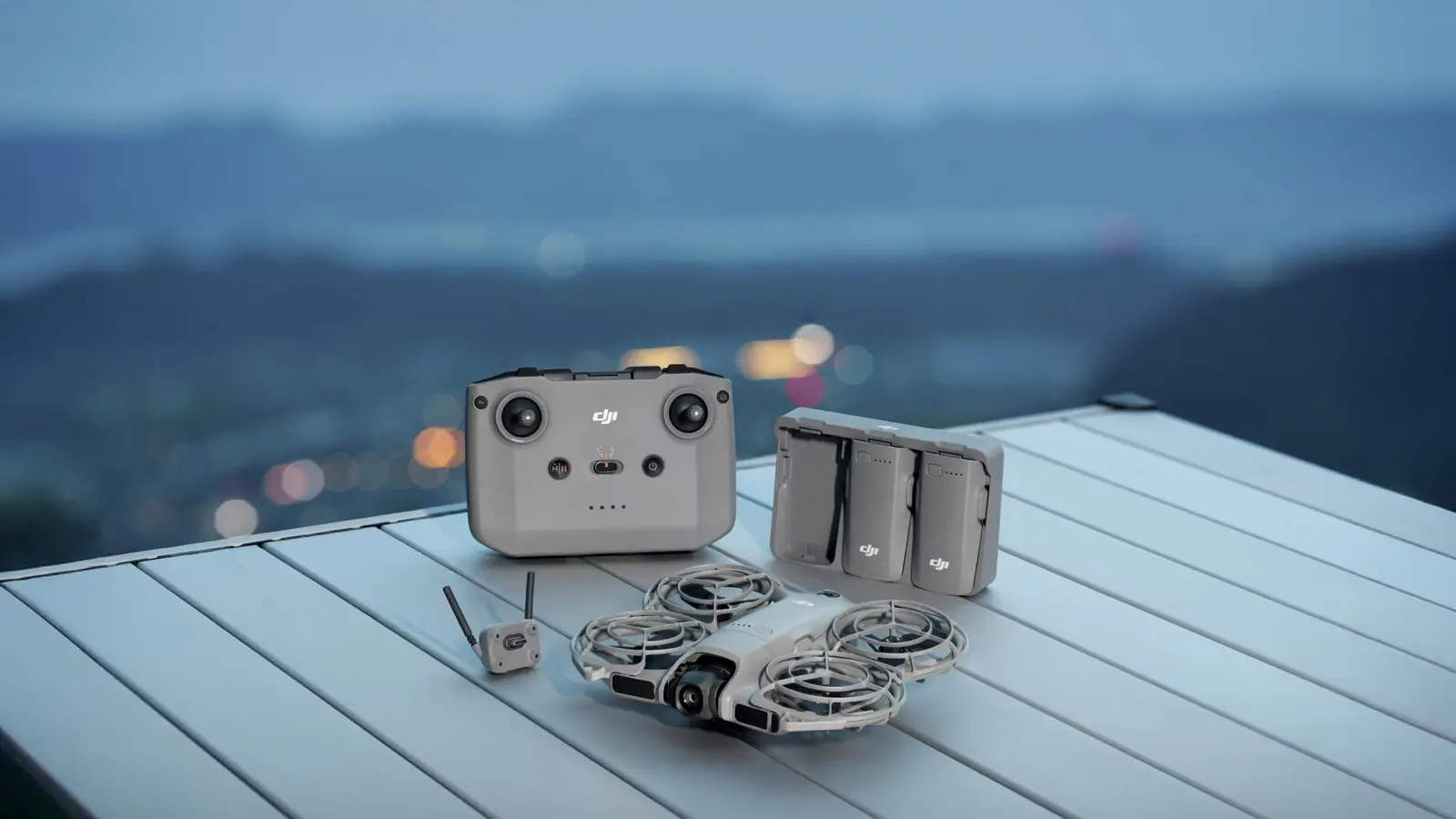 A gray DJI Neo 2 drone with four propeller guards on a table next to its small controller, a three-battery charging hub and a clip-on transceiver, with city lights behind.