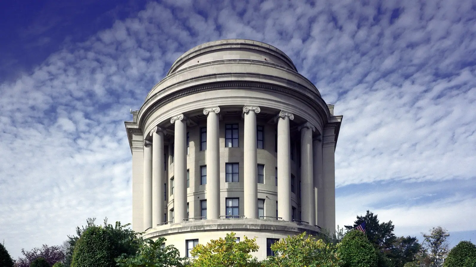 The round, columned Federal Trade Commission building in Washington, D.C., seen from a garden under a partly cloudy blue sky.