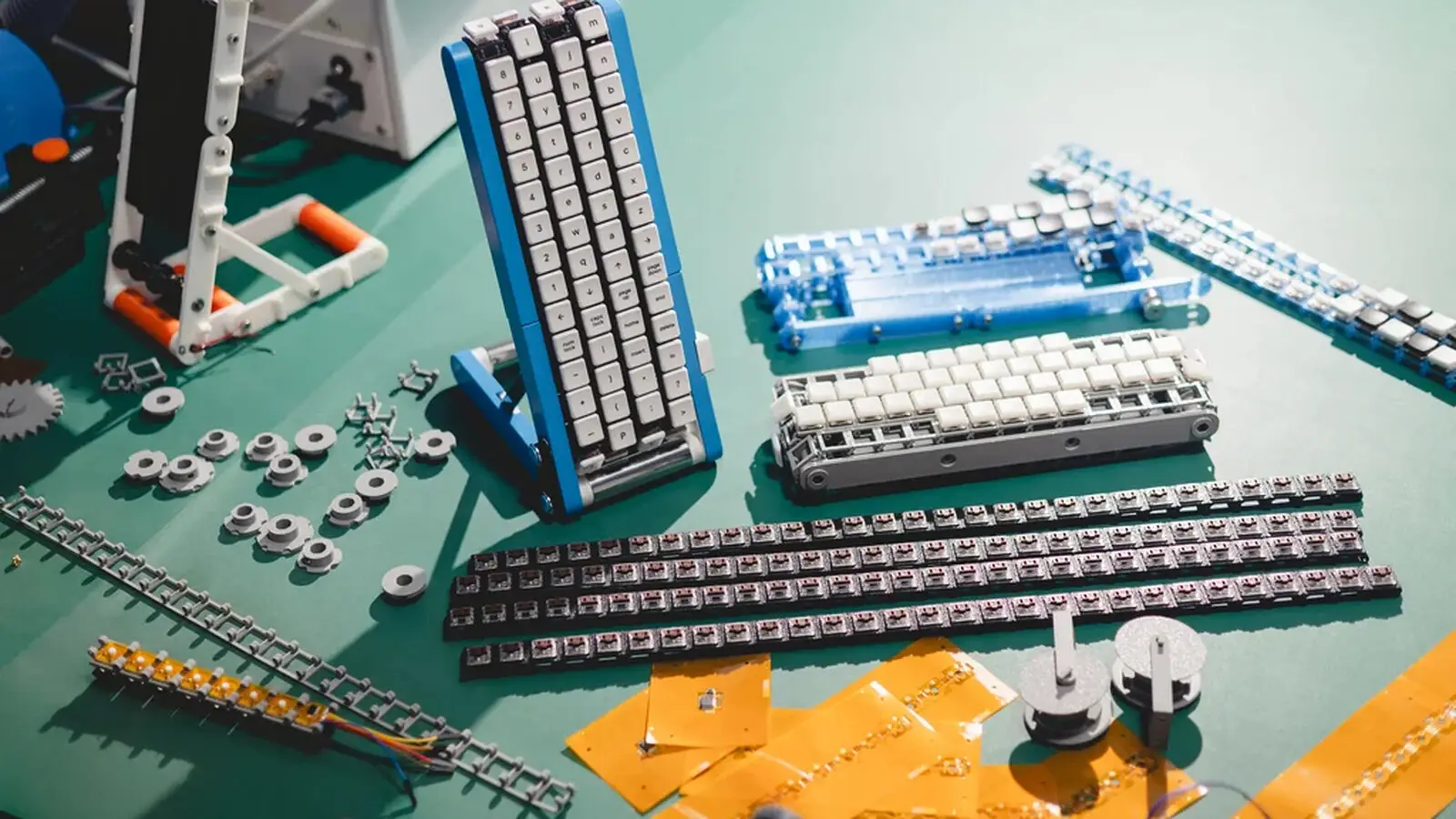 Parts of the Gboard conveyor belt keyboard laid out on a green table, with the assembled blue-framed keyboard standing upright among key belts, rollers and flex circuits.
