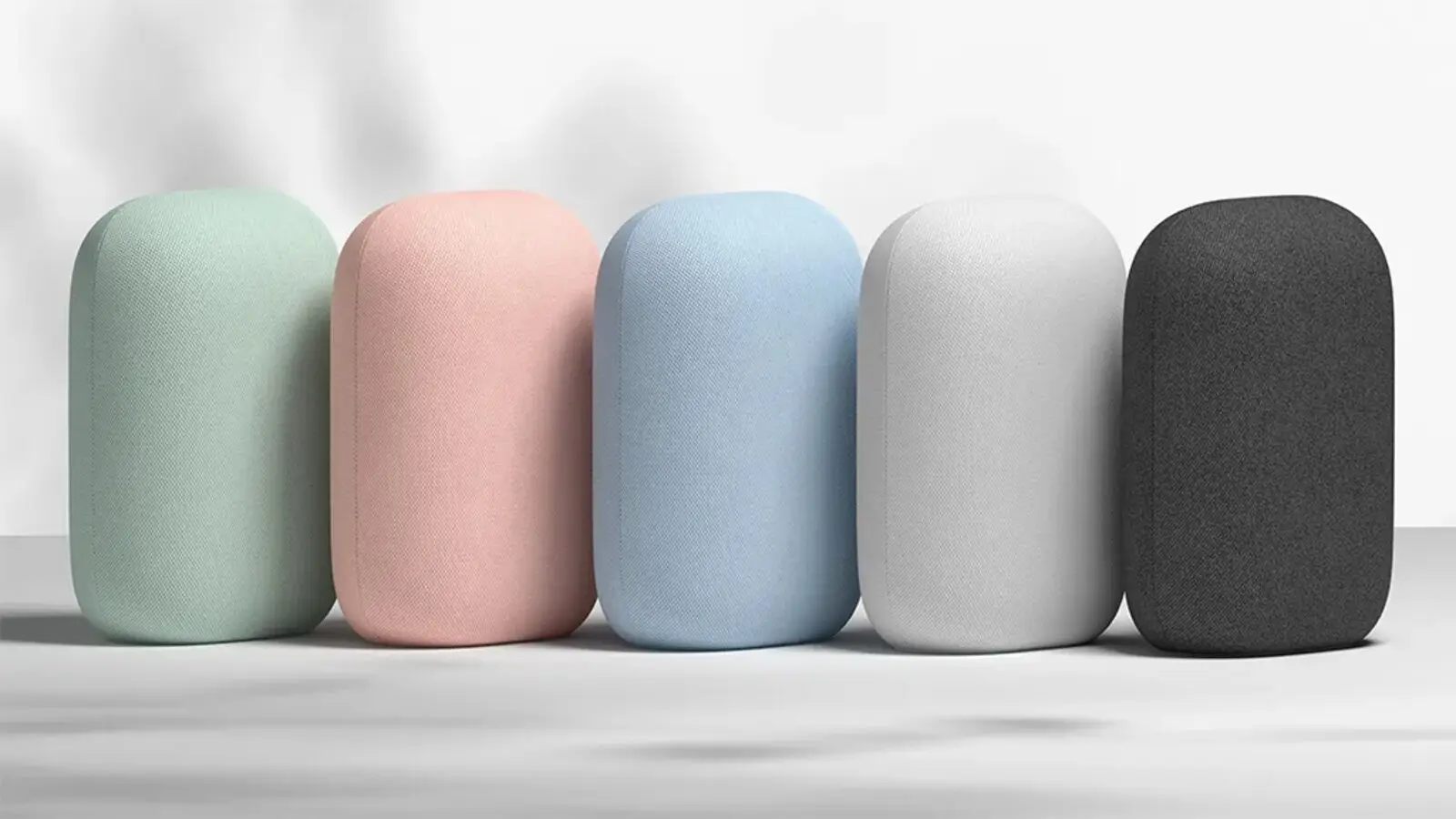 Five Google Nest Audio speakers in mint, pink, blue, white and charcoal fabric finishes, standing in a row against a pale background.