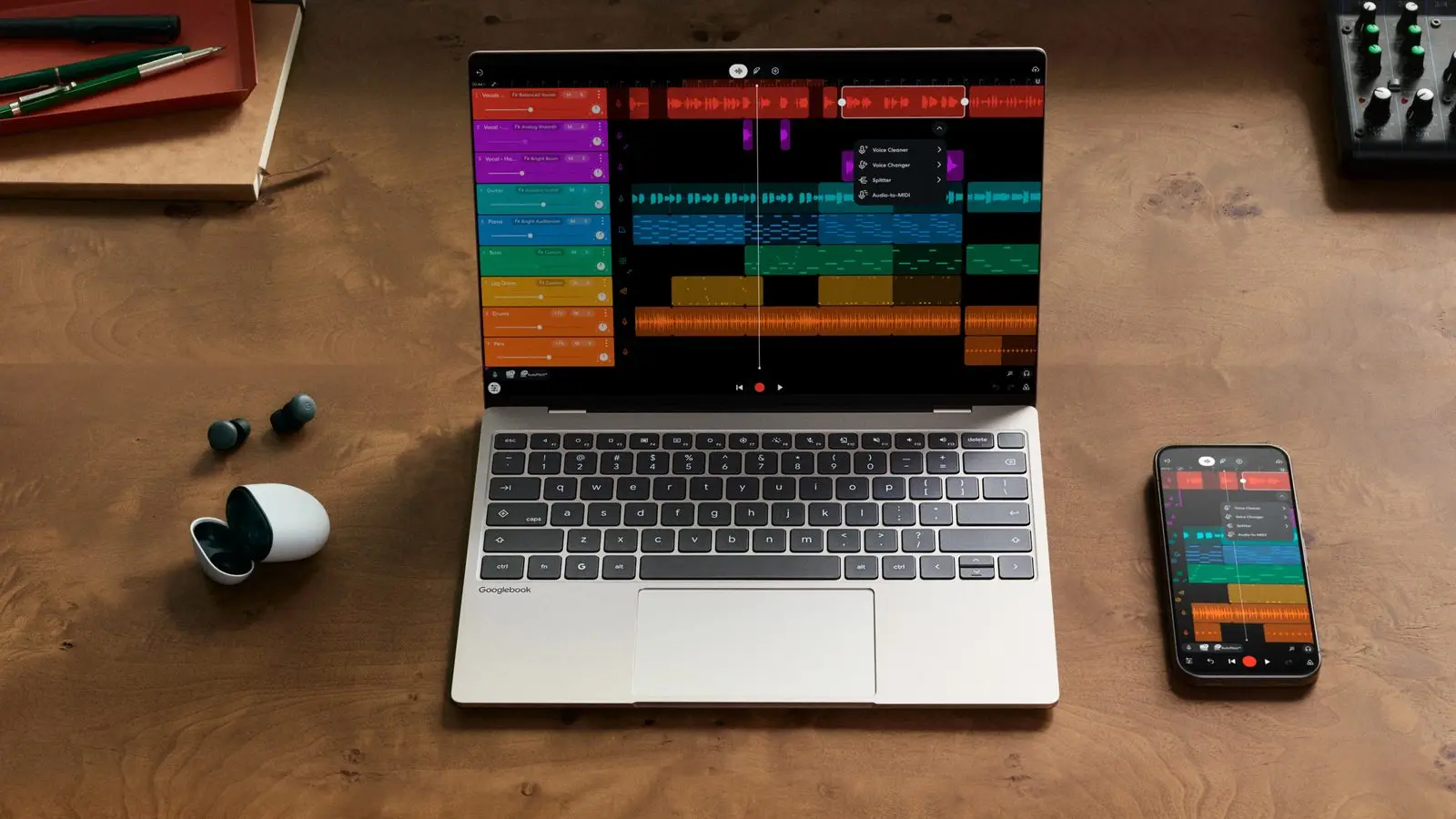Google's promotional photo of an open Googlebook on a wooden desk, with a music-editing app on its screen, a phone showing the same app and a pair of earbuds beside it. It is not the Chromebook in this story.