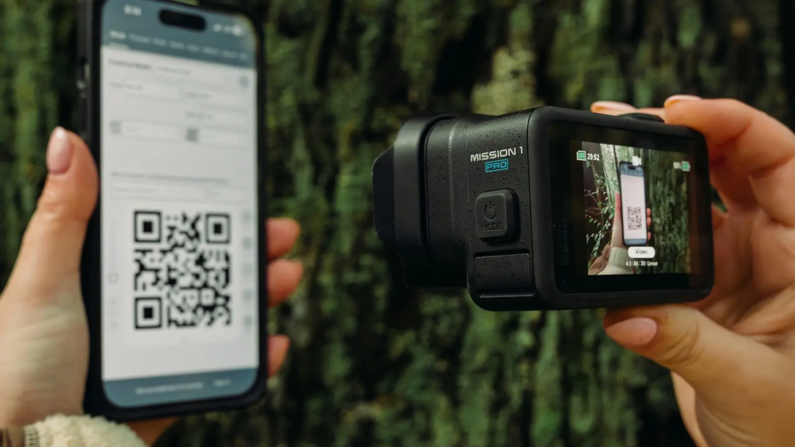 A hand holds a GoPro Mission 1 Pro camera, its rear screen showing a live view, beside a hand holding a phone that displays a QR code, against a mossy tree trunk.