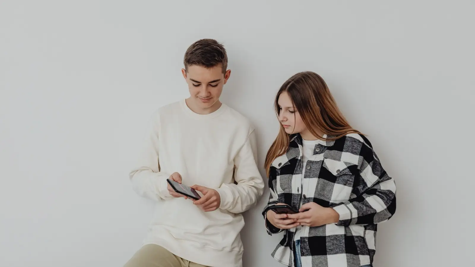 Two teenagers in casual clothes stand against a white wall, each looking down at a phone.