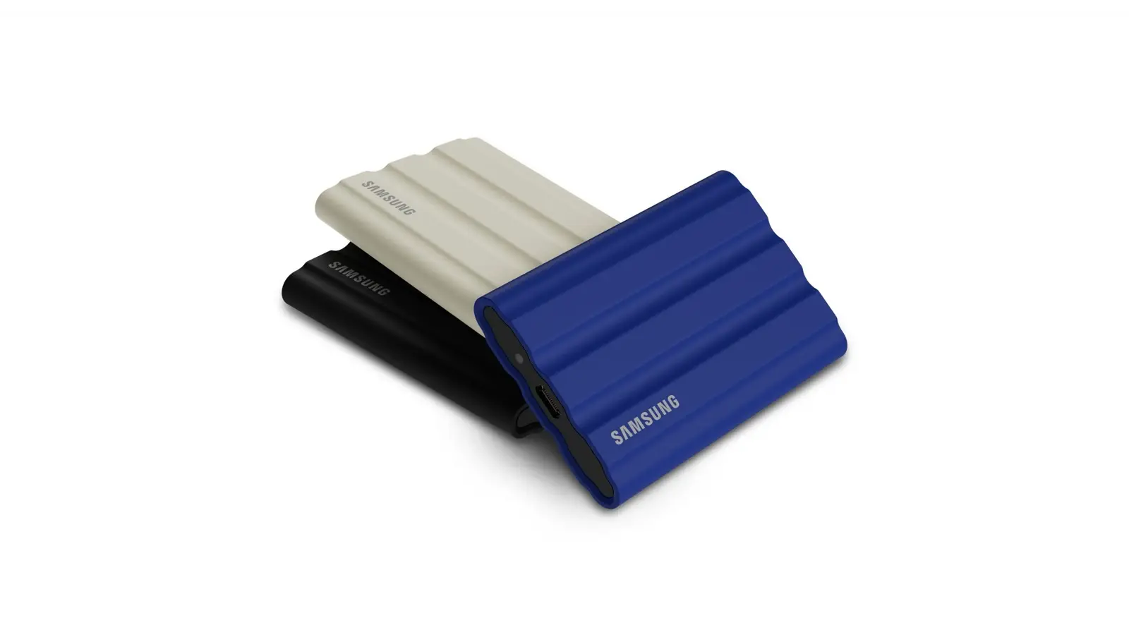 Three Samsung T7 Shield rugged portable SSDs stacked at an angle on white, in beige, black and blue, the blue one showing its USB-C port.