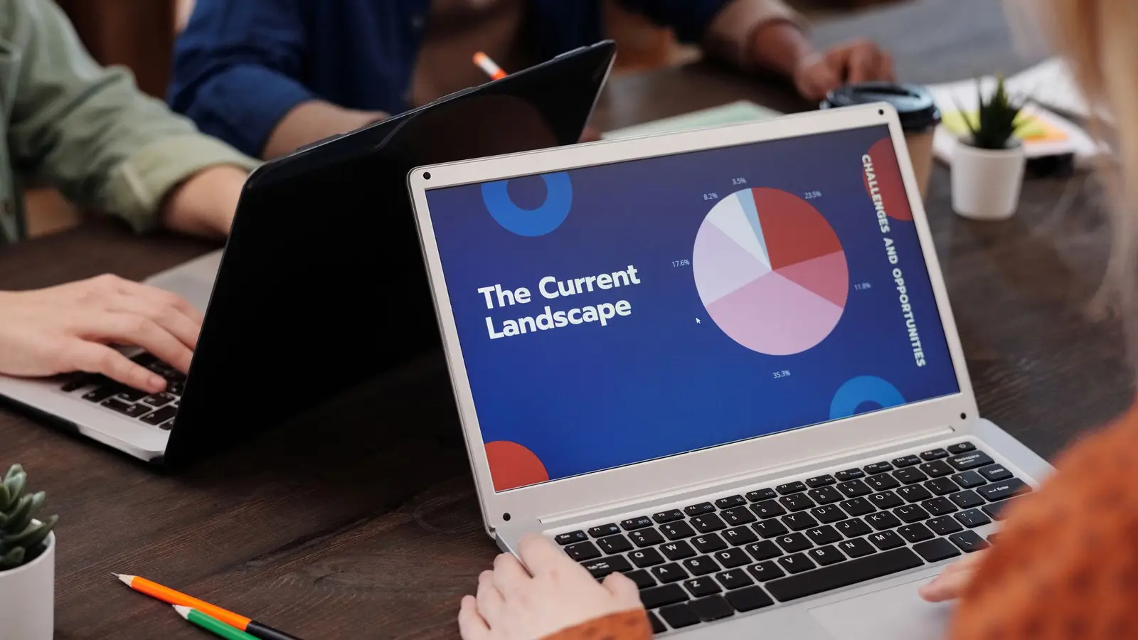 A laptop on a wooden desk shows a presentation slide with a pie chart while other people work on laptops around it.