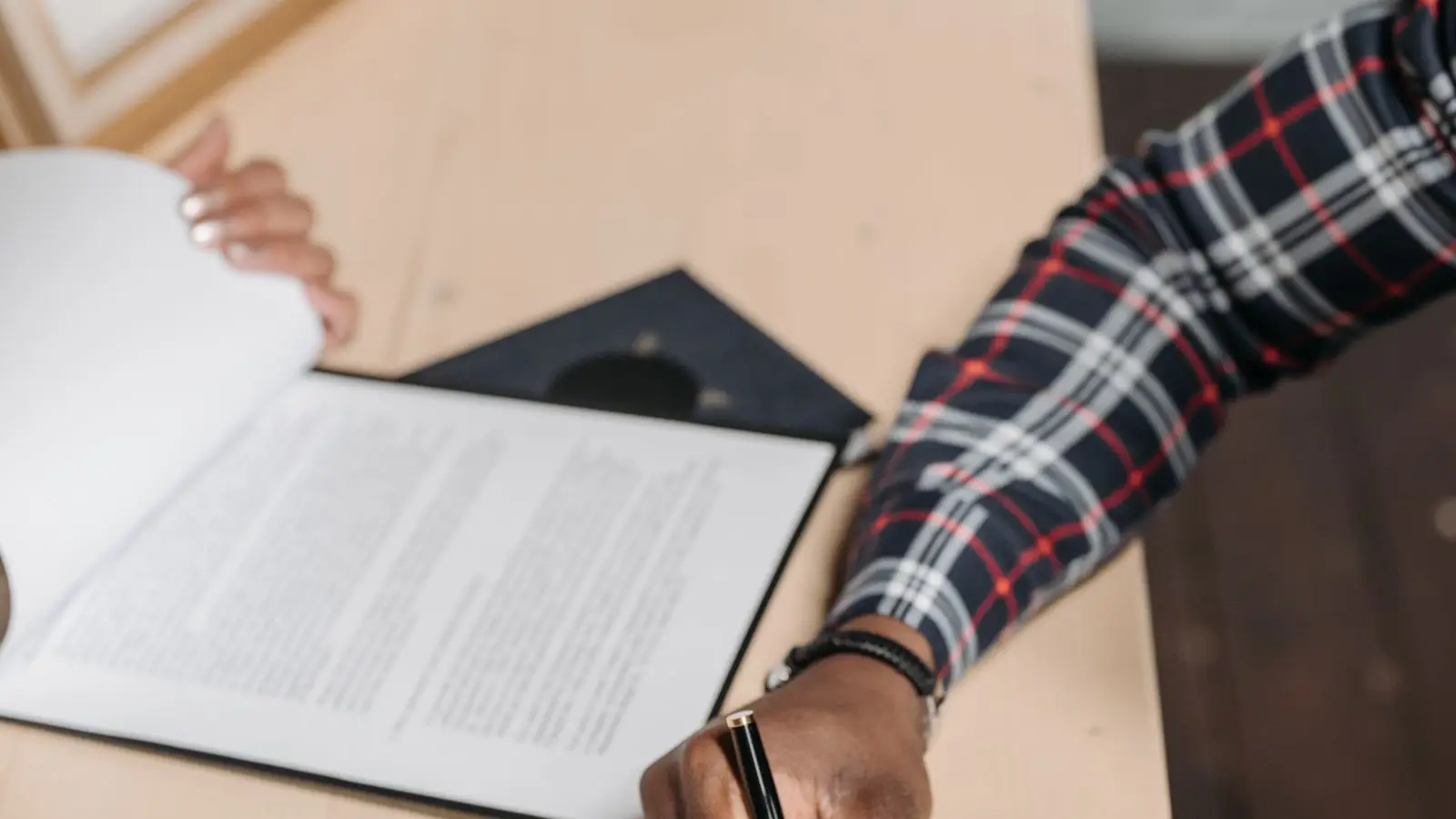 A hand signing a bound document with a black pen at a wooden desk.
