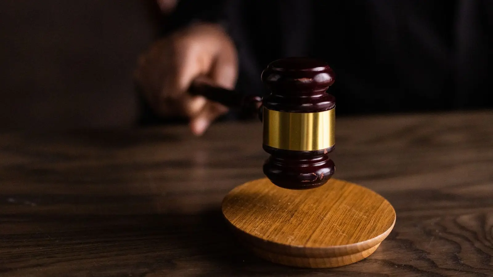 A wooden gavel raised above its sound block on a dark wooden desk, a hand holding it out of focus behind.