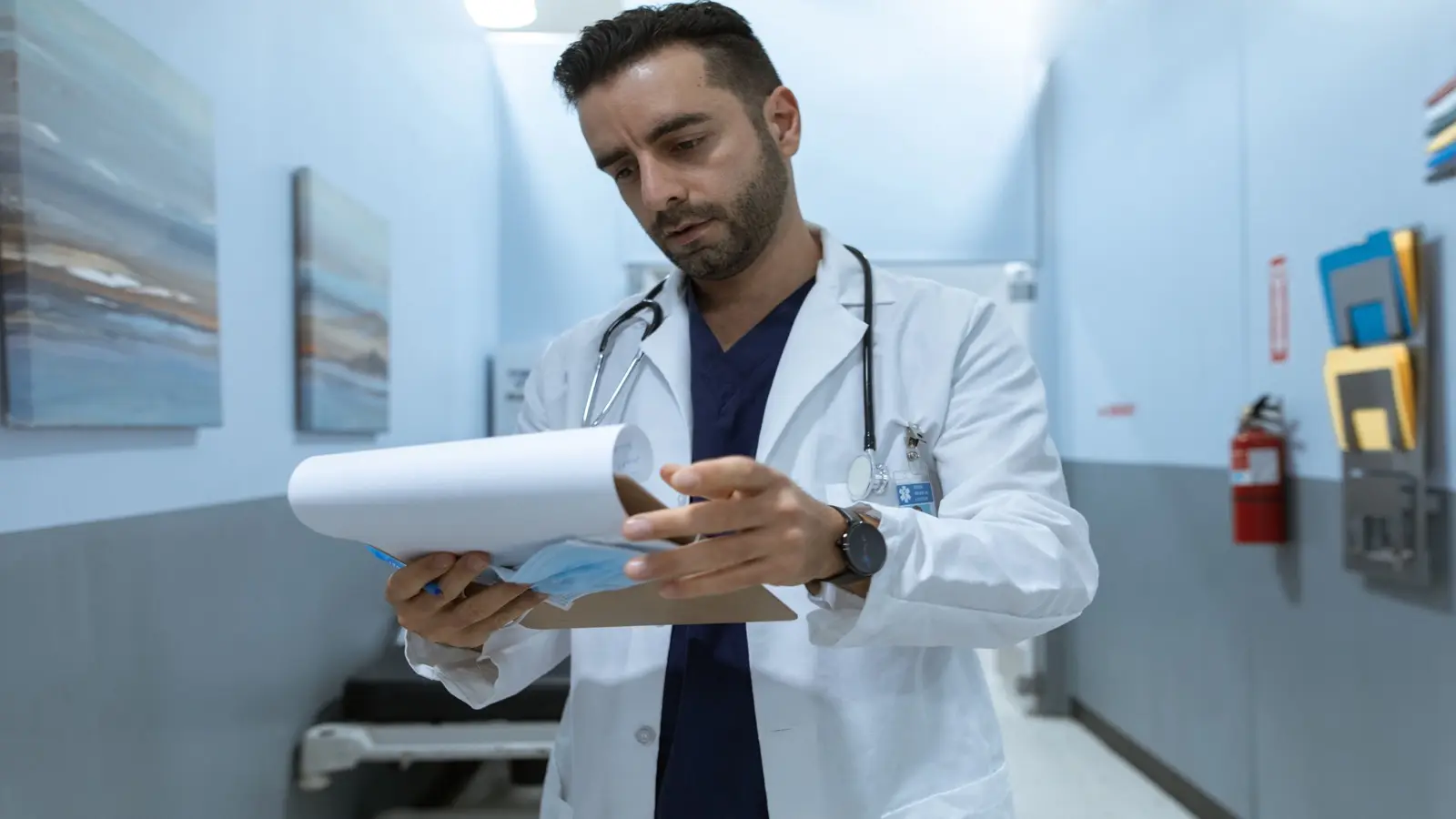 A doctor in a white coat and scrubs reads papers on a clipboard in a hospital corridor.