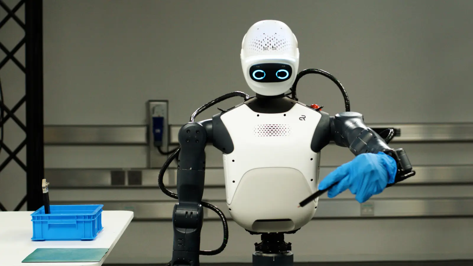 A white humanoid robot torso with glowing blue ring-shaped eyes, one hand in a blue glove holding a thin black tool, beside a blue crate on a white table.