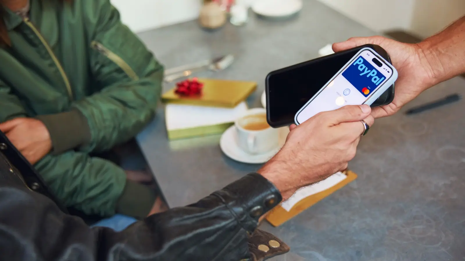 Two hands hold a phone showing a PayPal tap-to-pay screen against a card reader at a cafe table.