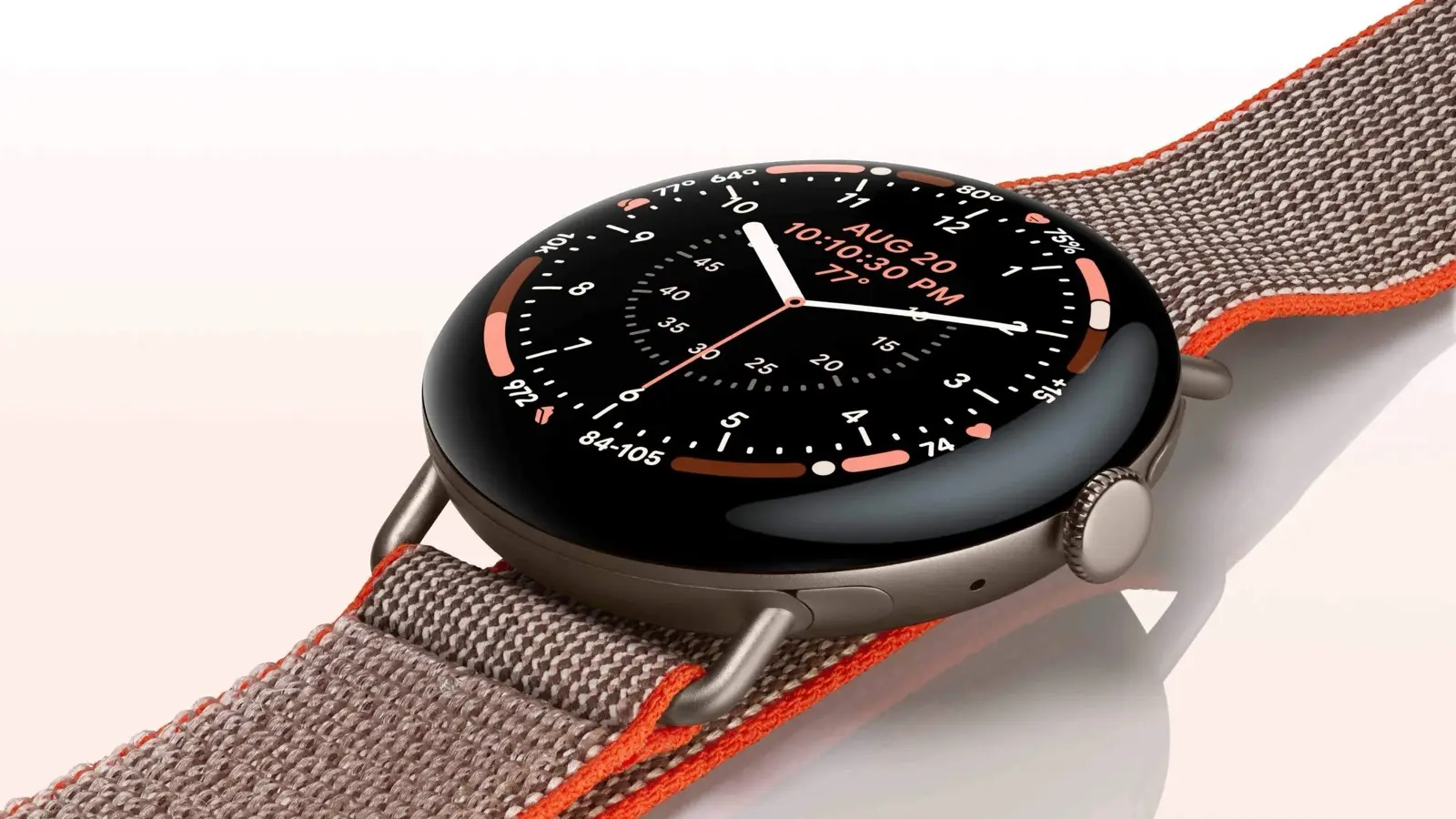 A round Pixel Watch with an analog watch face on a woven brown and orange band, against a pale background.