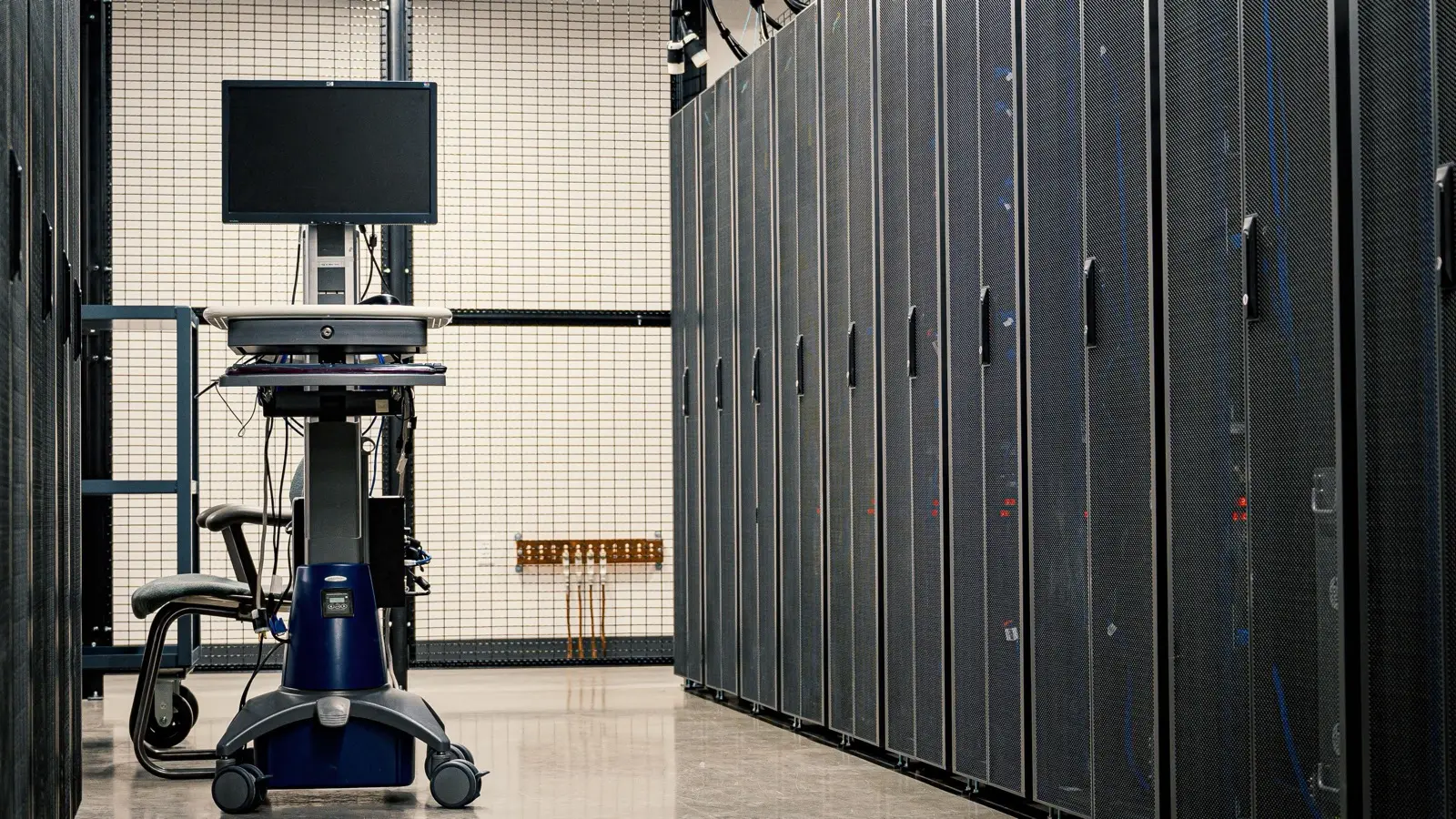 Rows of dark server racks along a data center aisle, with a monitor on a wheeled cart beside them.