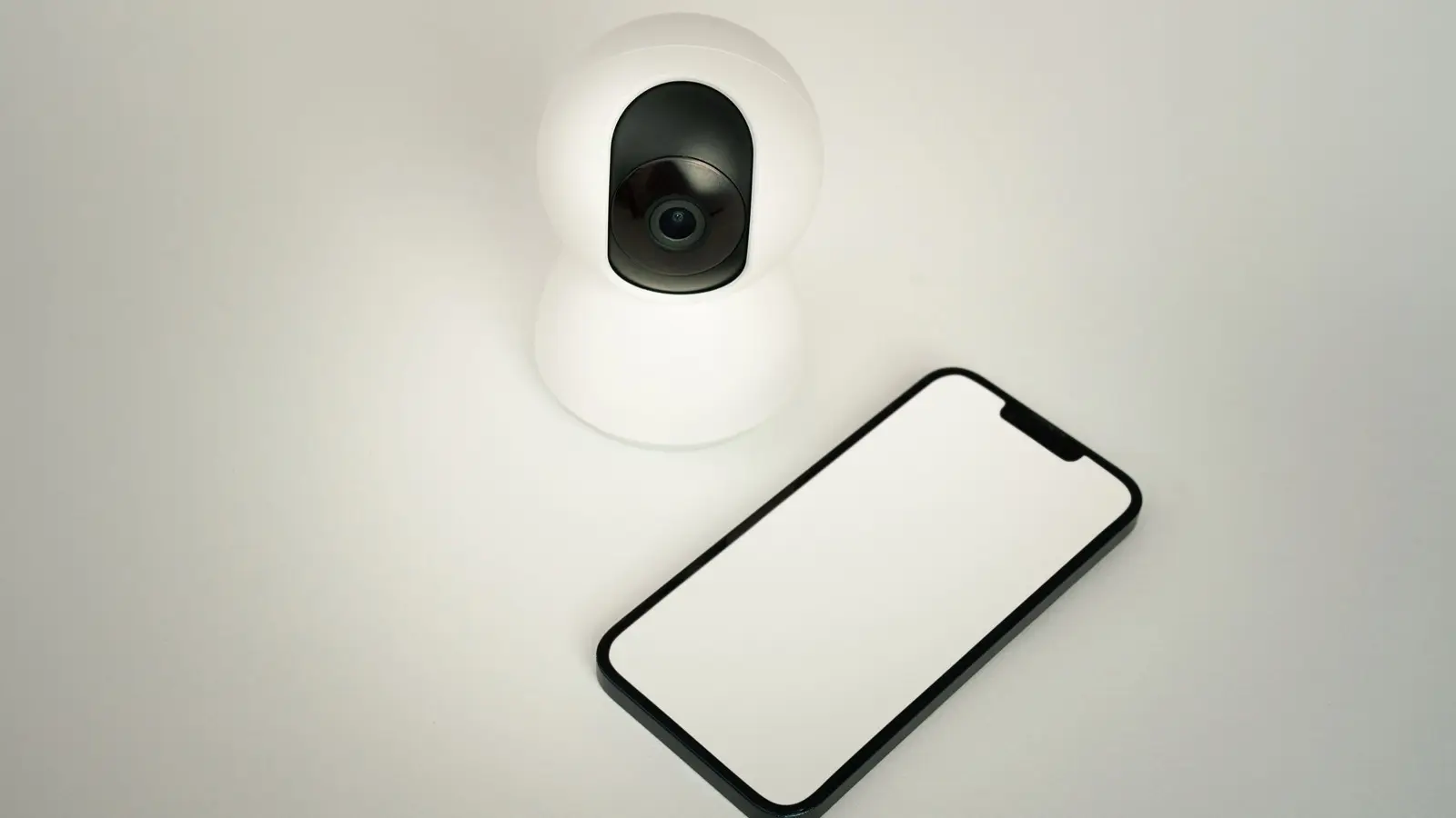 A white pan-tilt indoor security camera with a black lens window on a pale surface, next to a smartphone with a blank white screen.