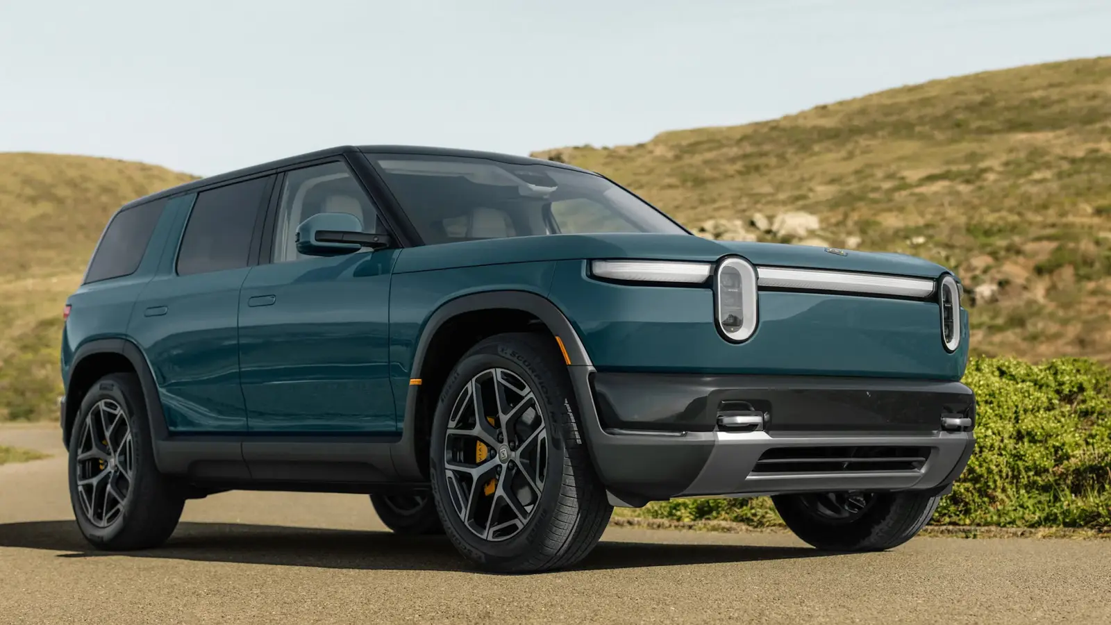 A teal Rivian R2 SUV parked on a paved pull-off in front of grassy hills under a pale sky.