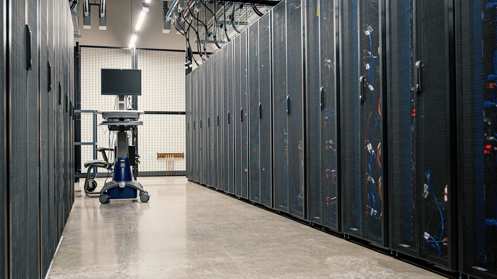 A long aisle of dark server racks in a data center, with cables running overhead.