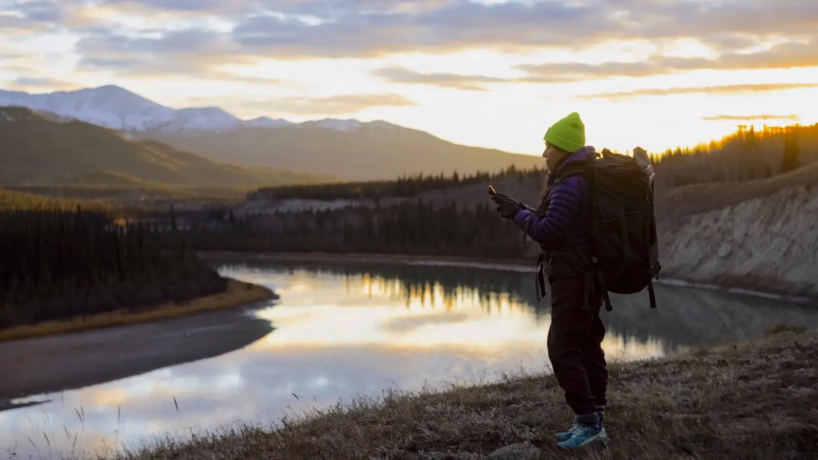 A hiker in a green hat checks her phone beside a river at sunset, with snowy mountains behind her.