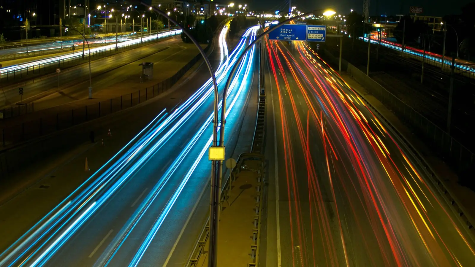 Long-exposure night photo of a multi-lane highway with blue and red streaks of car lights, street lamps and an overhead blue road sign.