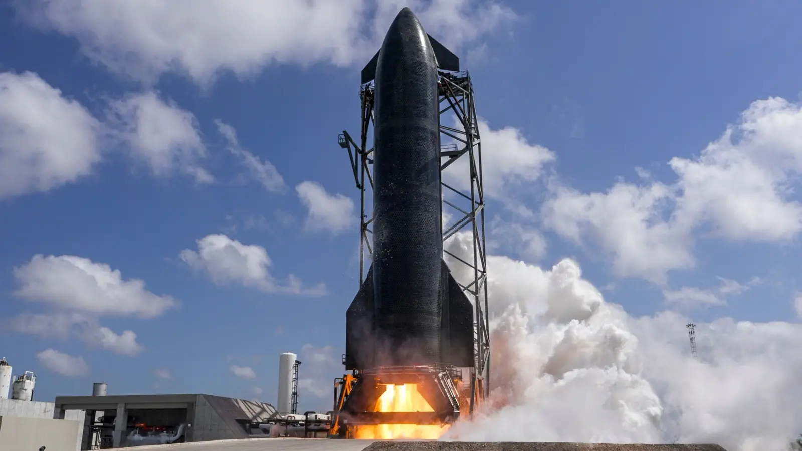 A Starship upper stage on a steel test stand during a static fire, with flame and white smoke at its base under a blue sky.