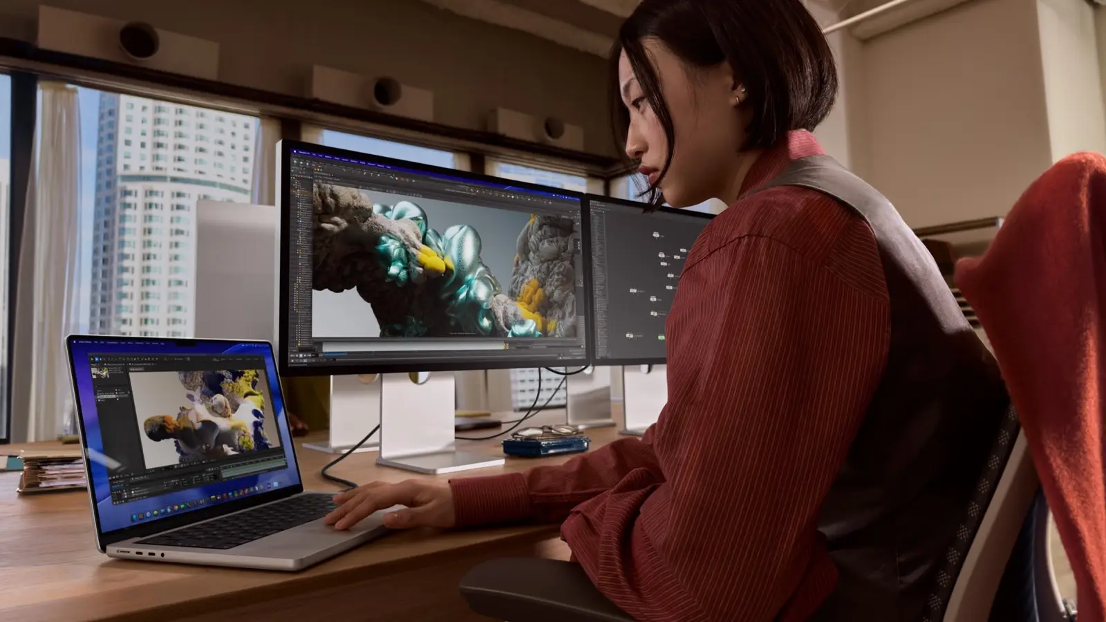 A creative professional works on a 3D animation on Apple's current 14-inch MacBook Pro next to two external displays, in an Apple press image. It is the current model, not the rumored OLED MacBook Pro.