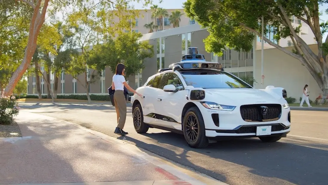 A white Waymo Jaguar I-Pace robotaxi with a roof-mounted sensor pod stopped at a tree-lined curb while a woman reaches for the rear door.